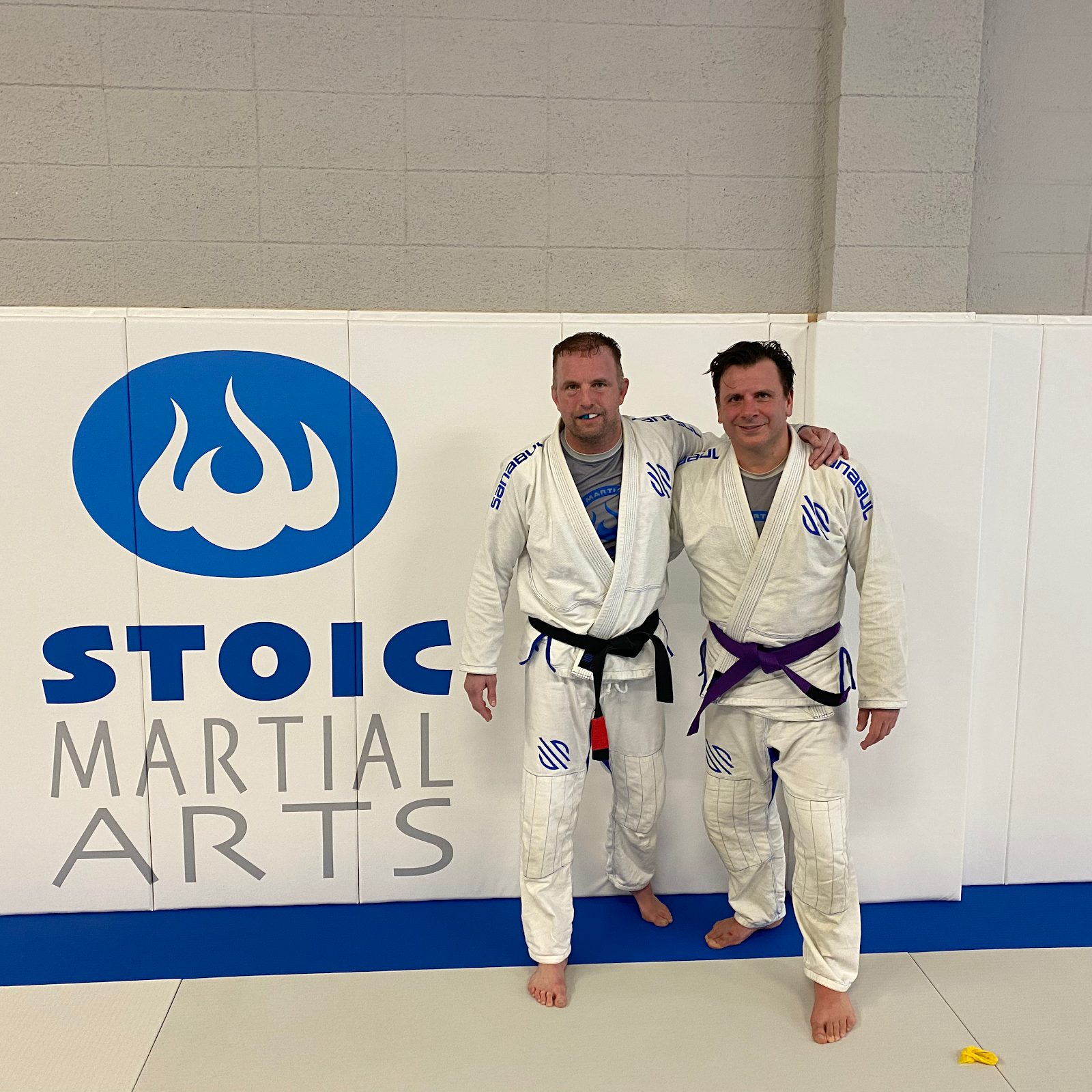 Adult BJJ students smiling in front of the Stoic Martial Arts logo wall in Chanhassen, Minnesota
